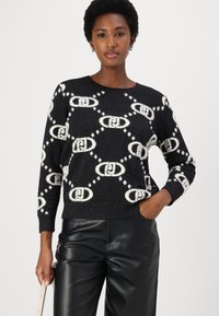 Zwarte trui met een wit kettingpatroon, ronde hals, lange mouwen en ribgebreide boorden. Draagt het met zwarte leren broek, handen in de zakken.