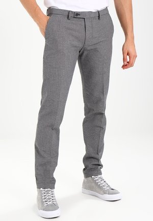Man met slim fit grijze, gestructureerde broek met voorzakken en grijze hoge sneakers met witte veters, staand met één hand in zijn zak.