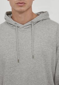 Sudadera gris claro hecha de tela texturizada con capucha ajustable, que presenta puntas metálicas en el cordón y un corte holgado.