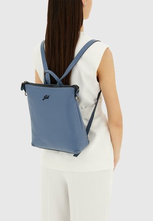 Donna con abbigliamento bianco senza maniche che indossa uno zaino in pelle blu con cerniera nera e logo sul retro, su uno sfondo semplice.