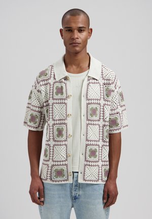 Mann trägt ein kurzärmeliges Häkelhemd mit floralen quadratischen Mustern über einem weißen, gerippten T-Shirt und hellblauen Jeans.