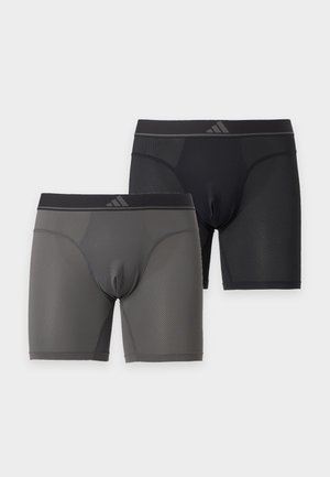 Homme grand, torse nu, à la peau foncée, debout pieds nus sur fond blanc, portant un boxer noir, les bras croisés et regardant sur le côté.