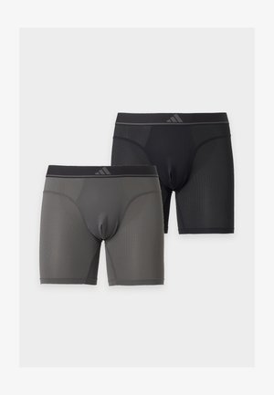 Ψηλός, γυμνόστηθος άντρας με σκουρόχρωμο δέρμα στέκεται ξυπόλητος μπροστά σε λευκό φόντο, φοράει μαύρα μπόξερ και έχει τα χέρια σταυρωμένα κοιτώντας προς τα πλάγια.