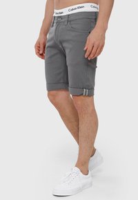 Grå denimshorts med opfoldede manchetter, featuring en knaplukning og bæltestropper. Båret med hvide sneakers. Calvin Klein-linningen er synlig.