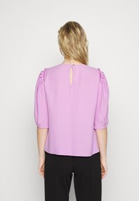 Blusa lilla con dettaglio a buco chiave sulla schiena, maniche corte a sbuffo e pieghe sottili. Realizzata in tessuto morbido, abbinata a pantaloni neri.