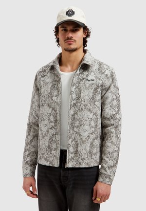 Man wearing a gray patterned zip-up jacket, white shirt, black pants, white cap with black logo, and gold rings on fingers, facing forward.