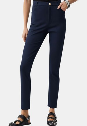 Donna che indossa pantaloni slim fit blu navy con tasche frontali e bottone dorato, abbinati a sandali neri borchiati e bracciali argentati.