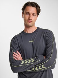 Mand med mørkt hår iført en mørkegrå langærmet trøje med grønne kilsstriber på ærmerne og "hummel performance" logo på brystet.