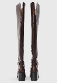 Hohe dunkelbraune Lederstiefel mit Blockabsätzen und Knöchelriemen, aufrecht stehend, von hinten auf einem weißen Hintergrund gezeigt.