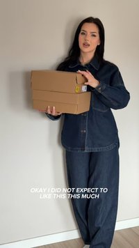 Jeune femme en tenue en jean foncé tenant deux boîtes à chaussures en carton empilées contre un mur uni avec un texte exprimant surprise et plaisir.