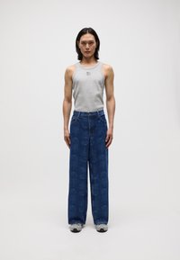 Graues Tanktop mit Bärenmotiv, kombiniert mit weit geschnittenen dunkelblauen Jeans mit floralem Muster. Das Model trägt graue Sneakers.