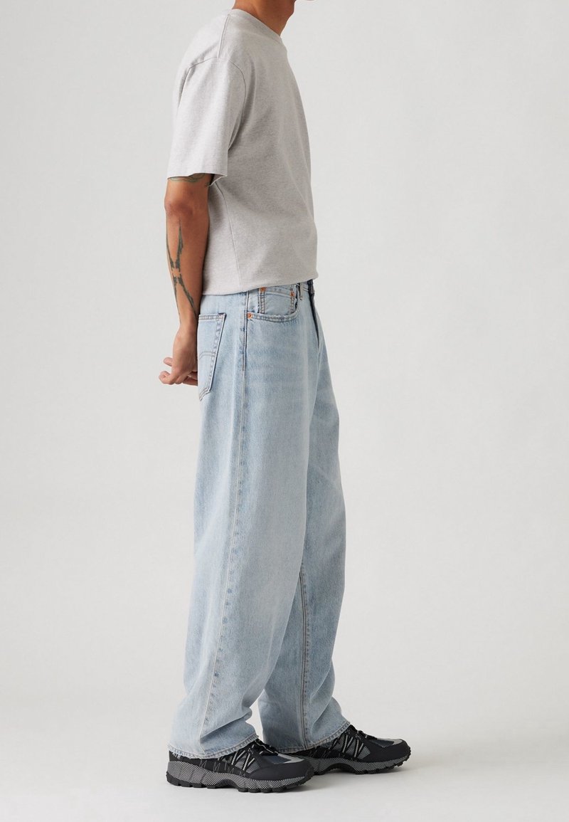 Homme portant un t-shirt gris clair, un jean bleu clair ample et des baskets noires, debout de profil les mains croisées derrière le dos, sur un fond uni.