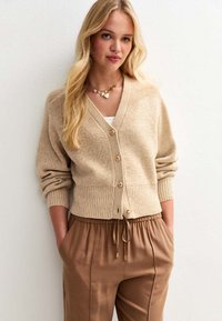 Beige stickad cardigan med djup V-ringning, långa ärmar, ribbade mudd och knäppning med knappar. Matchad med ljusbruna byxor med dragsko.