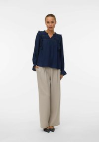 Blusa blu navy con rouches, scollo a V e maniche lunghe, abbinata a pantaloni beige larghi e scarpe a punta con pois.