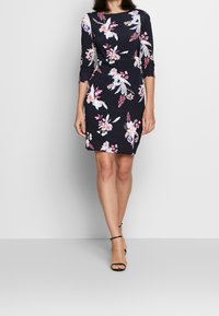 Nainen, joka on pukeutunut mustaan, vartaloa myötäilevään mekkoon, jossa on vaaleanpunaisia ja valkoisia kukkakuoseja sekä kolme neljäsosaa hihasta, yhdistettynä mustiin nilkkasandaaleihin.