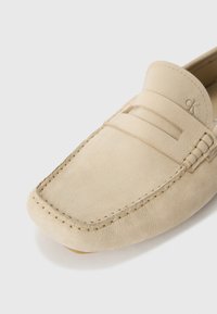 Beige leather loafers with stitched detailing, featuring a flat heel and a decorative strap across the vamp. Subtle logo on the side.