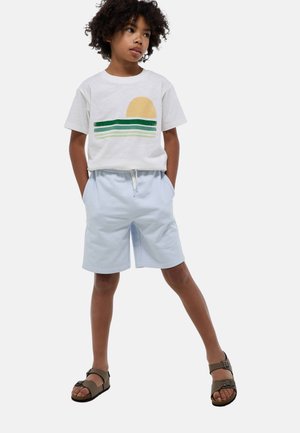 T-shirt blanche avec un motif de soleil jaune et des rayures vertes, associée à un short bleu clair avec une taille élastique et un cordon de serrage. Sandales marron.