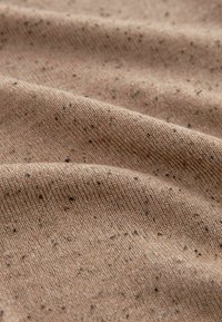 Gros plan sur un tissu beige doux avec de petites taches sombres et une texture tricotée visible, montrant des plis et des ondulations délicats.