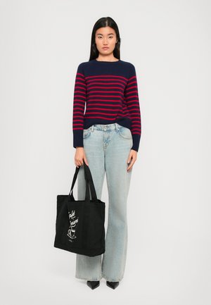Mørkeblå og rødstripet genser med lange ermer, ledsaget av lyseblå, vide jeans. Modellen holder en svart tote-bag med hvit tekst og grafikk.