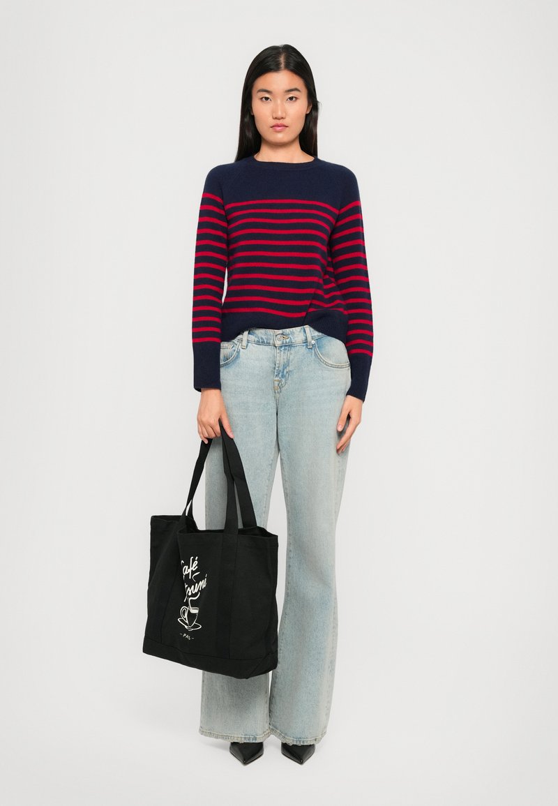 Mørkeblå og rødstripet genser med lange ermer, ledsaget av lyseblå, vide jeans. Modellen holder en svart tote-bag med hvit tekst og grafikk.