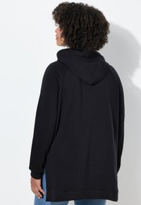 Persona con capelli ricci che indossa un ampio felpa nera e jeans blu, rivolta di spalle contro uno sfondo bianco semplice.