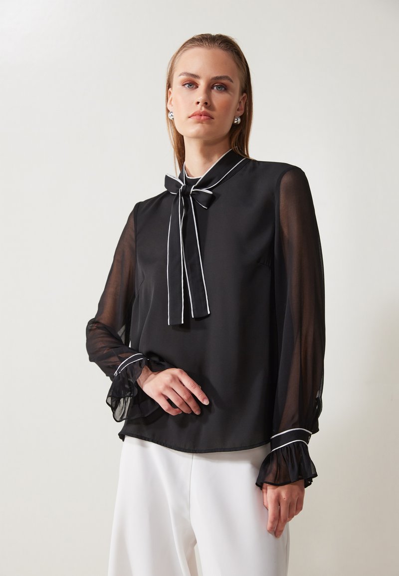 Blusa nera con maniche lunghe trasparenti e collo con fiocco. Presenta dettagli in bianco sui polsini e sul colletto. Vestibilità ampia con una texture fluida.