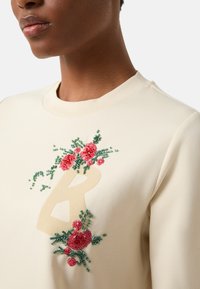 Krémově zbarvená mikina s květinovým motivem, vyšívaným červenou a zelenou nití, a velkým béžovým písmenem "B". Hladká textura látky.