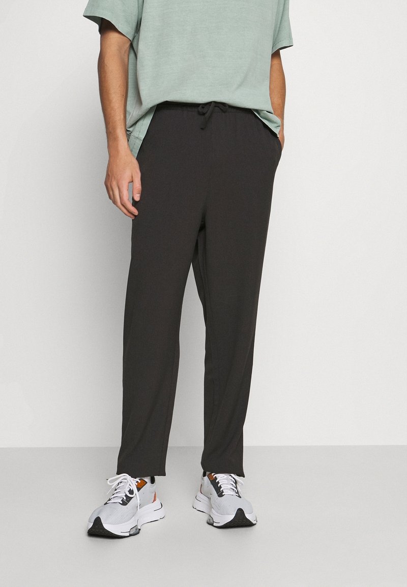 Pantalon foncé à coupe ample avec une taille à cordon de serrage, des poches et une texture lisse, associé à une chemise à manches courtes vert clair et des baskets blanches.