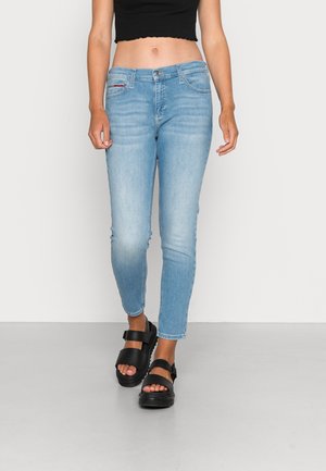 Femme portant un jean skinny bleu clair, des sandales noires à larges brides, et un top noir court, marchant devant un arrière-plan clair uni.