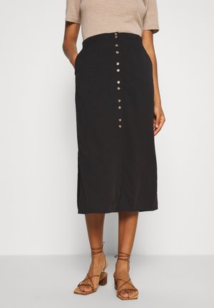 Femme portant une jupe midi noire à boutons sur le devant, un haut beige à manches courtes et des sandales marron à talons épais et lanières, debout les mains dans les poches.