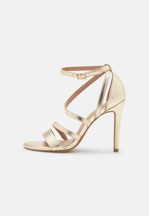Gouden metallic hoge hakken sandaal met meerdere kruisende bandjes, open teen en enkelband met een gespsluiting. Soepele textuur.