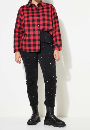 Personne portant une chemise à carreaux rouge et noire, un pantalon noir avec des détails en perles blanches et des bottes épaisses noires, debout devant un fond blanc.