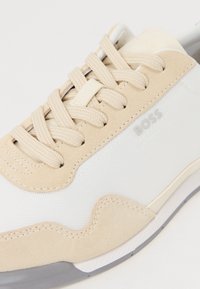 Beige semsket og hvit mesh-sneaker med avrundede kanter, snøringdesign og subtil merkevaretrykk på siden. Gummisåle.