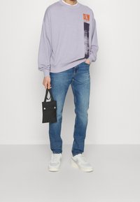 Person som bär en ljuslila sweatshirt med Calvin Klein-logga, blå jeans, vita sneakers och håller en liten svart handväska.