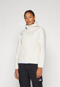 Giacca impermeabile beige chiaro con cappuccio, polsini elasticizzati, due tasche frontali e logo sul petto sinistro. Finitura in tessuto liscia e resistente.