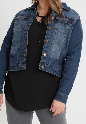 Frau trägt eine kurze blaue Jeansjacke über einer schwarzen Bluse und grauen Hosen und steht vor einem einfarbigen Hintergrund.