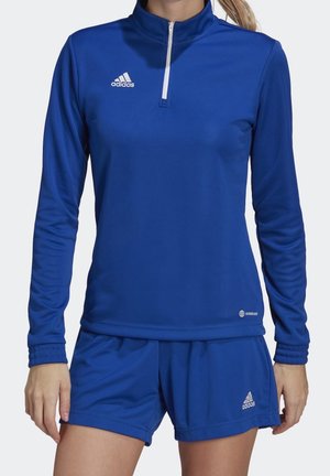 Kvinde iført en blå Adidas langærmet zip-up top og matchende blå shorts med Adidas-logoer på begge beklædningsgenstande.