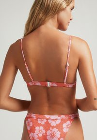 Conjunto de bikini en color coral con estampado floral en rosa; cuenta con tirantes delgados ajustables, cierre de gancho en la parte posterior y un acabado sin costuras.