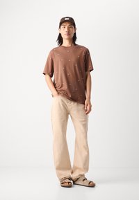 Brun bomulds T-shirt med små broderede designs, parret med lysebeige afslappede bukser og brune slidssandaler. Enkel kasket på hovedet.