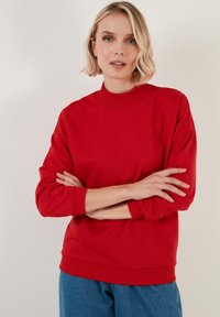 Sudadera roja con cuello alto, mangas largas y puños acanalados. La tela parece suave y el diseño es de corte holgado.