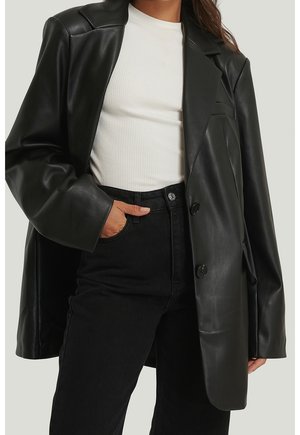 Femme portant un blazer oversize en similicuir noir, un top blanc côtelé et un jean noir taille haute, une main dans la poche.