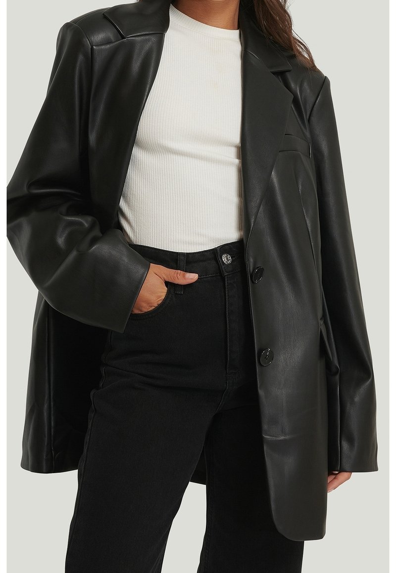 Femme portant un blazer oversize en similicuir noir, un top blanc côtelé et un jean noir taille haute, une main dans la poche.