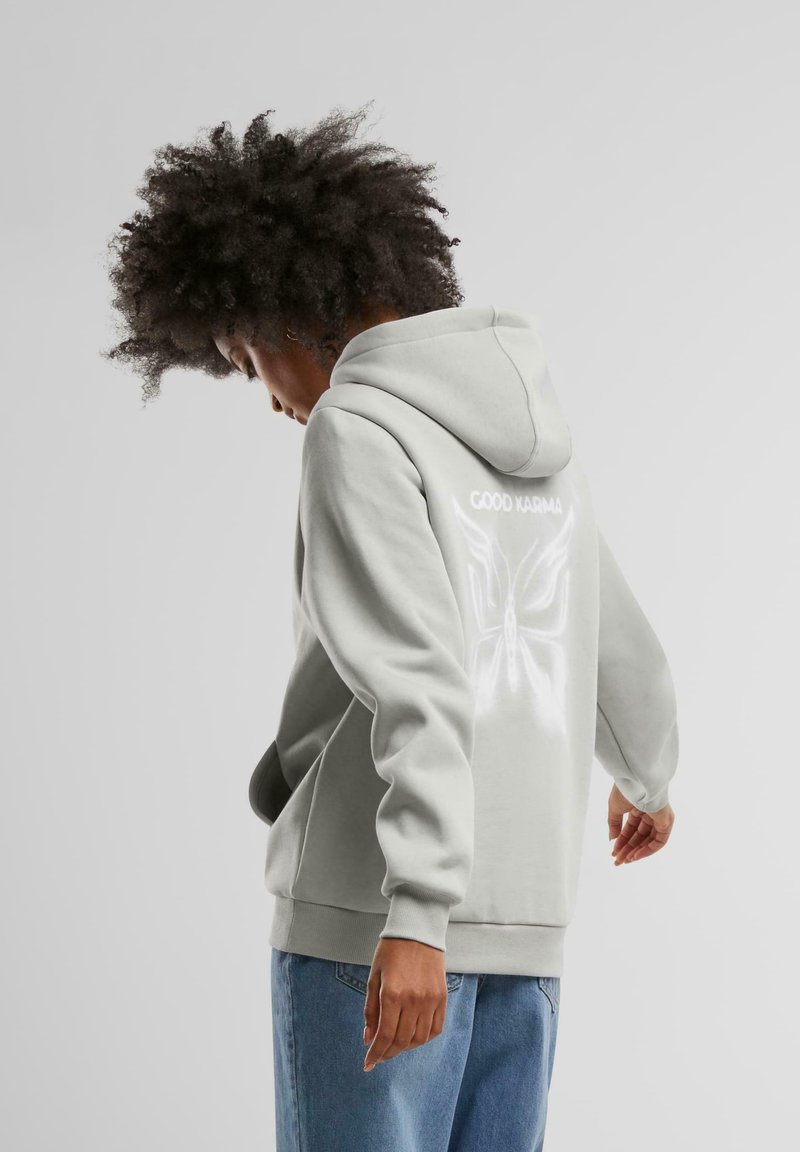 Ljusgrå hoodie med en rymlig huva, med en reflekterande fjärilsdesign och texten "GOOD KARMA" på baksidan. Mjuk textur.