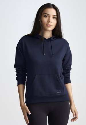 Young woman with long dark hair wearing a navy blue hoodie with front pocket and black leggings, standing against a plain background.