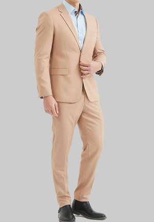 Costume beige avec blazer à simple boutonnage et pantalon assorti. Chemise bleue claire en dessous avec un motif à carreaux au col. Bottes noires.