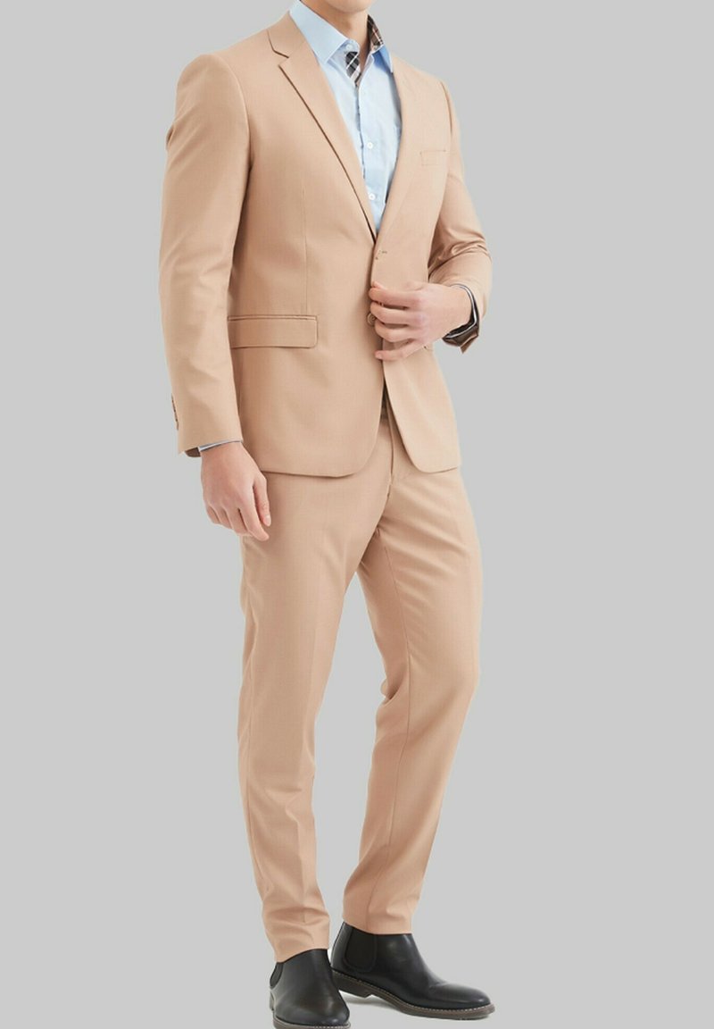 Costume beige avec blazer à simple boutonnage et pantalon assorti. Chemise bleue claire en dessous avec un motif à carreaux au col. Bottes noires.