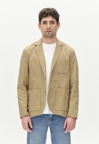 Beige, könnyű blézer két elülső zsebbel, puha anyagból, lehajtható gallérral és laza szabással, fehér póló és kék farmer fölött viselve.