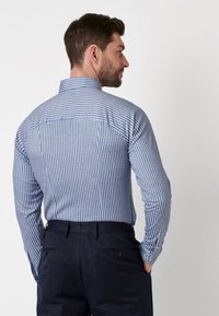 Man met donker haar in een blauw-witte verticaal gestreepte shirt en donkere broek, staand met handen in de zakken, naar rechts gericht tegen een witte achtergrond.