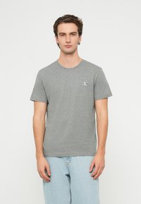 Šedé bavlnené tričko s krátkymi rukávmi, okrúhlym výstrihom a malým bielym logom na ľavej strane hrudníka. Hladká textúra, voľný strih.