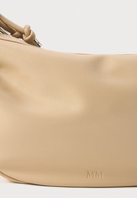 Beige nahkakaelakott, millel on sile tekstuur ja õrn sära, sisaldab embosseeritud "MM" logo ja ümarat disaini.